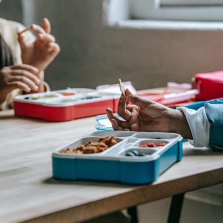 Crop anonymous classmates sitting at wooden table with lunch boxes and eating homemade food